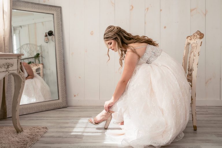 Gorgeous young bride sitting down in her wedding dress putting on her high heels in front of a mirror