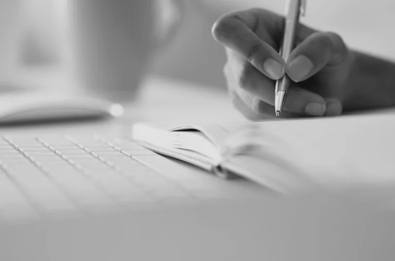 Black and white photo of pen writing in a notebook with a keyboard in front