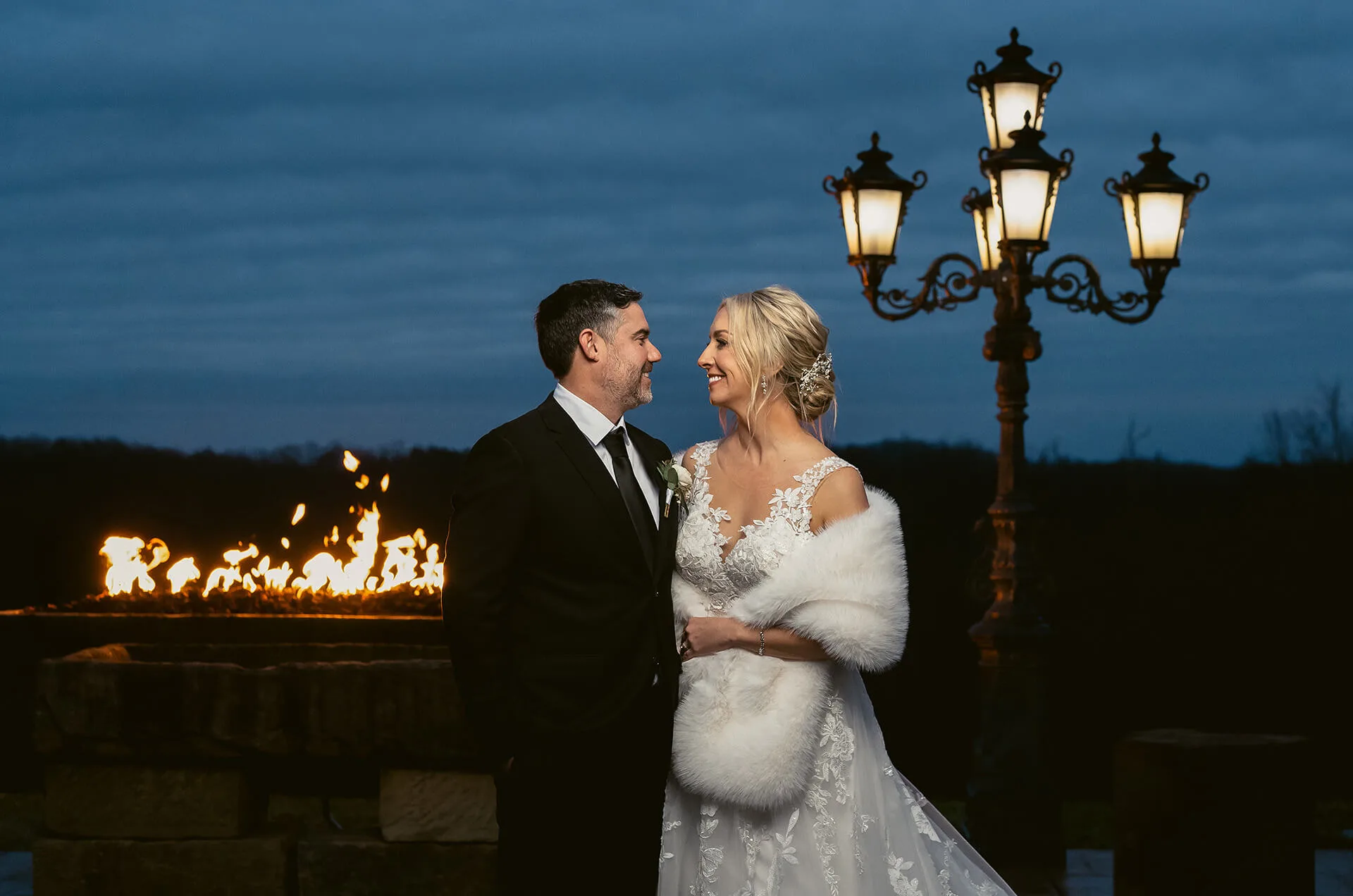 Night photograph of husband and wife in their wedding attire looking into each others eyes with fire burning behind them and a lamppost in the background